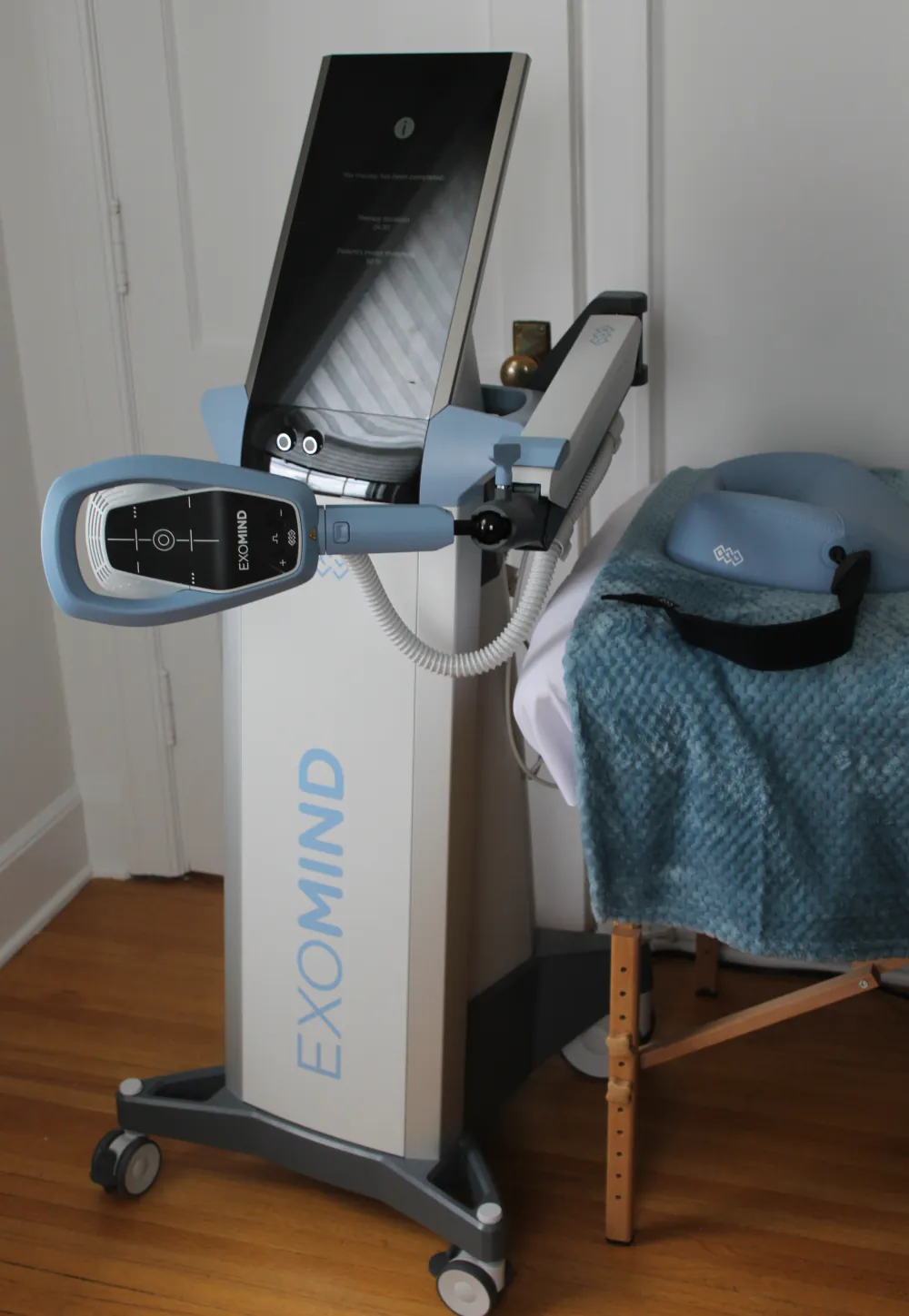 The EXOMind unit at the Centre — a screen on a mobile stand with an articulated arm and head-shaped coil, beside a treatment table with a blue blanket.