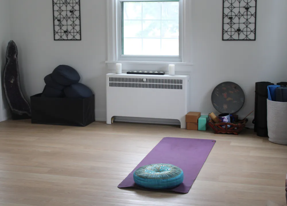A movement and meditation room with a yoga mat and cushion on a hardwood floor, plants by the window.