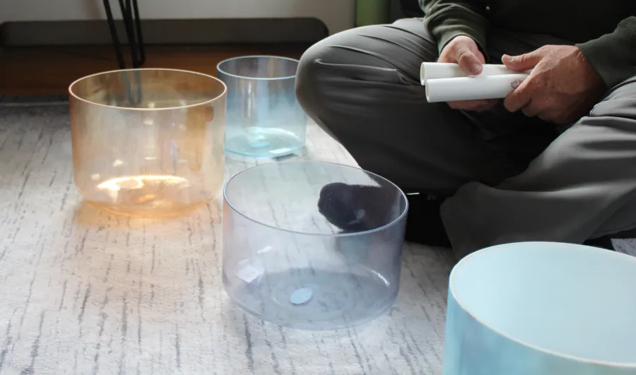 A practitioner sits cross-legged with a striker beside a set of crystal singing bowls, warm light across a hardwood floor.
